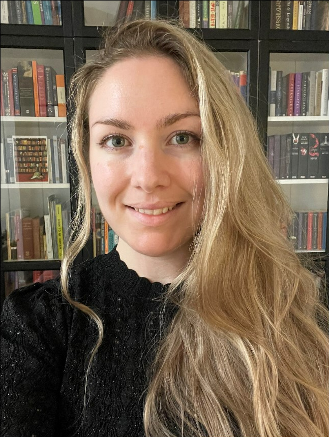 Head and shoulders photo of Ash Barber, smiling in front of bookcases. Ash is a Caucasian woman with long blonde hair and is wearing a black knit jumper.